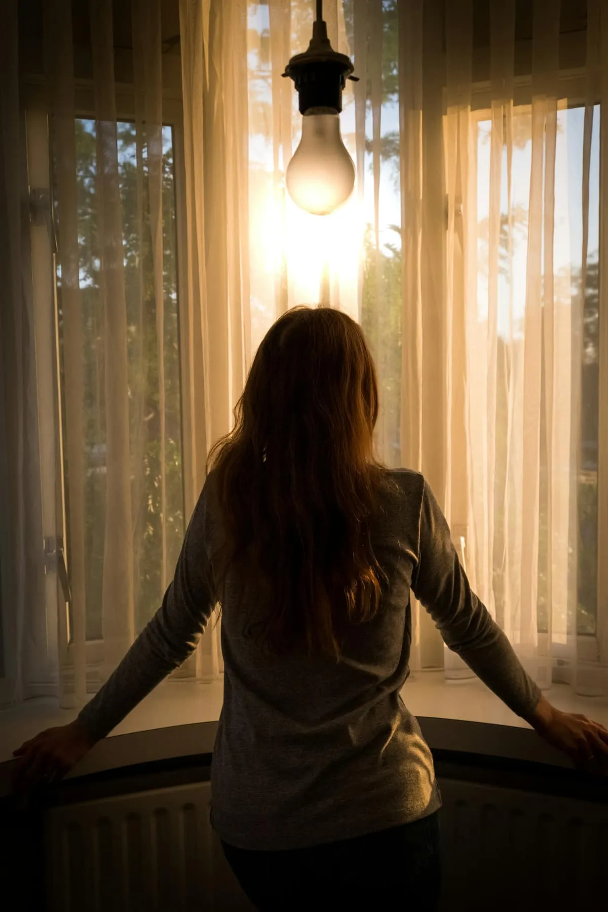 A young person standing alone by a window, backlit in soft natural light, a contemplative silhouette.