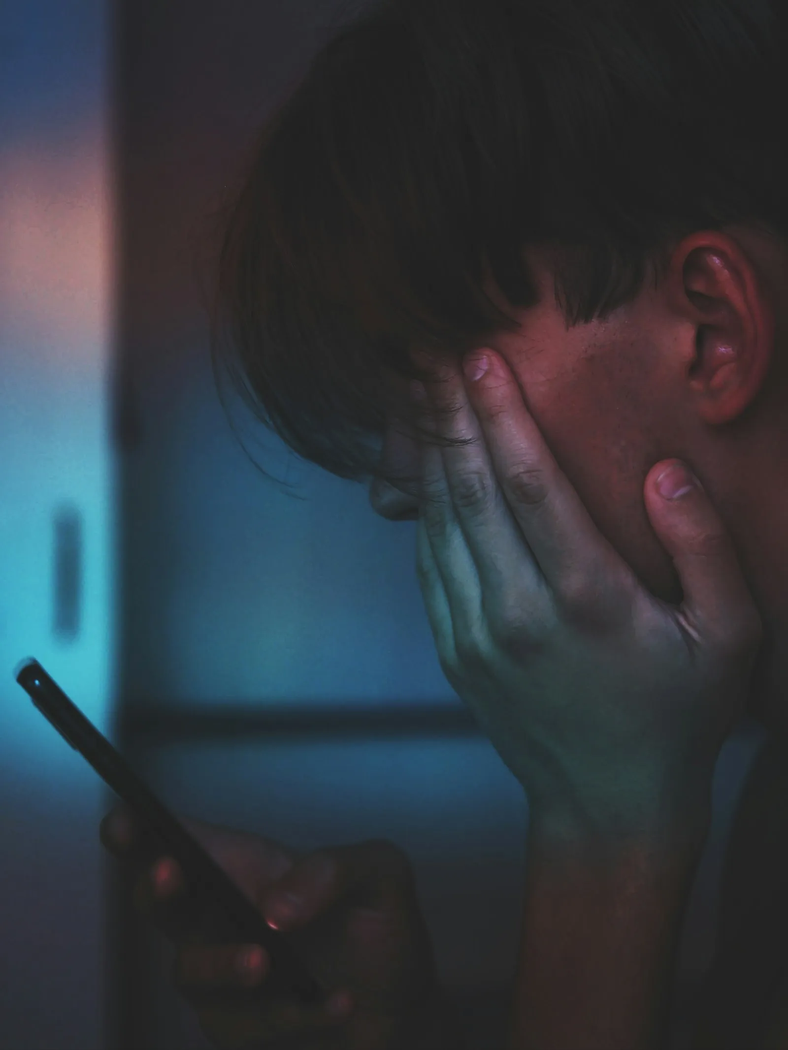 A glowing phone screen casting blue light on a teen's face, eyes downcast